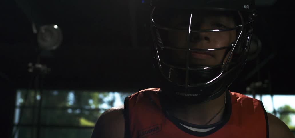 A young lacrosse player wearing a helmet stares intensely forward in a dramatically lit gym in slow motion