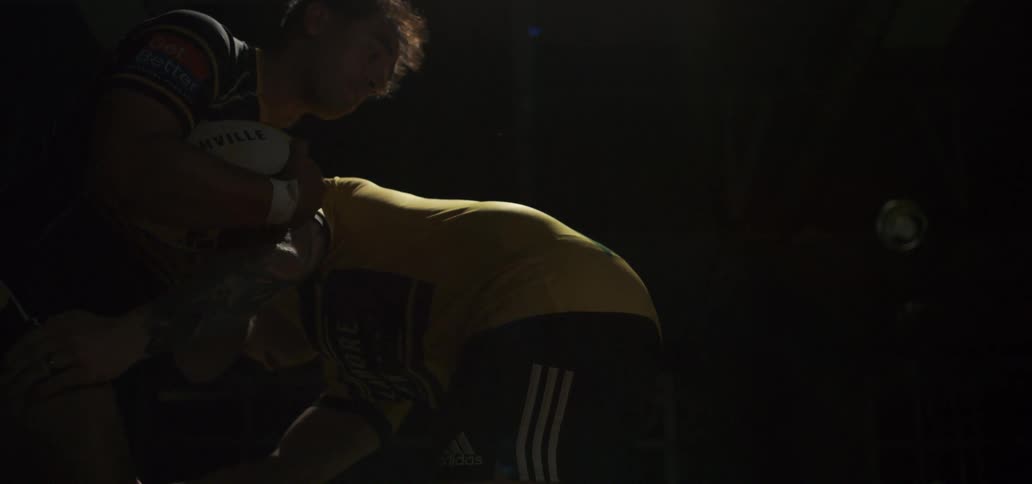 Two rugby players engage in an intense scrum in a dramatically lit, dark room in slow motion