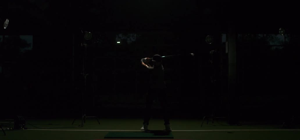 A man practices his powerful golf swing in slow motion inside a dramatically lit studio driving range