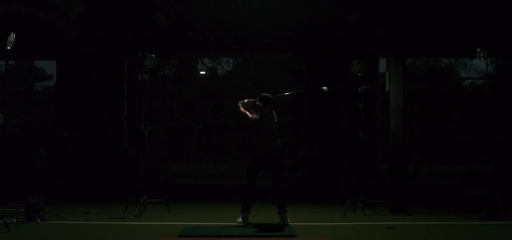 A male golfer in a dark indoor facility takes a powerful swing at a golf ball in super slow motion, dramatically lit by a flash