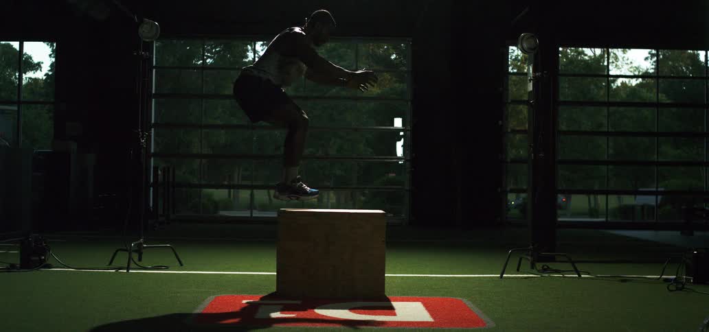 An athletic man lands a box jump in slow motion, highlighted by a powerful strobe light in a gym