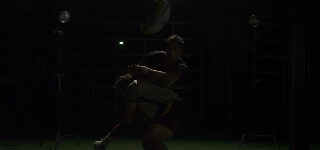 Two rugby players tackle each other in super slow motion under dramatic studio lights, sending the ball flying