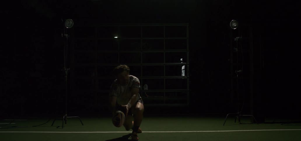A rugby player passes a ball in dramatic slow motion under spotlights inside a dark training facility