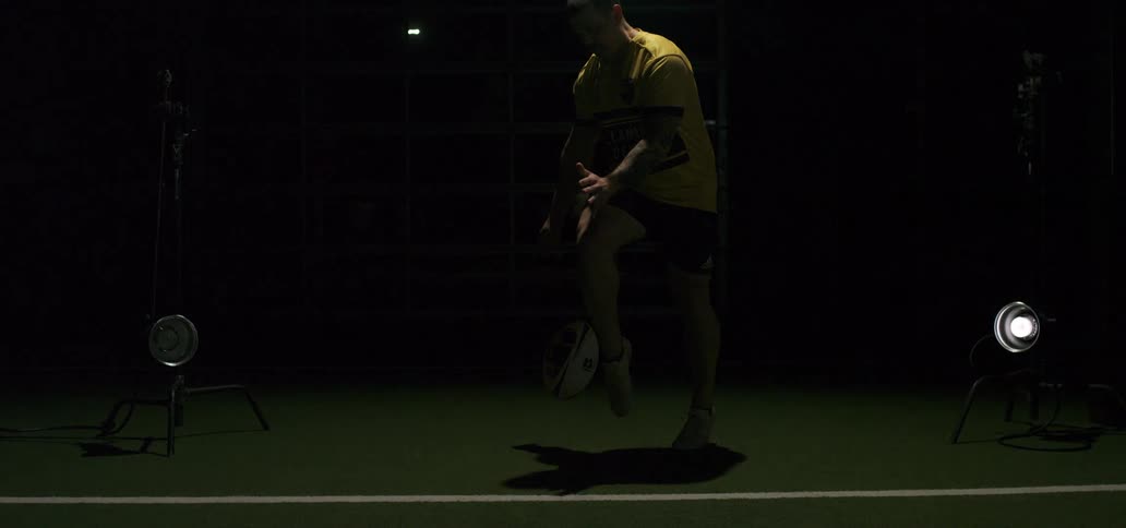 Focused rugby player executes a dropkick in a dramatically lit indoor facility, captured in extreme slow motion
