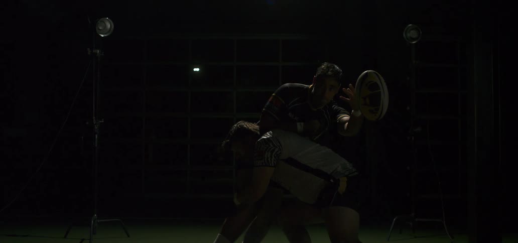 A powerful rugby player tackles his opponent, who is holding the ball, in dramatic slow motion under studio lights