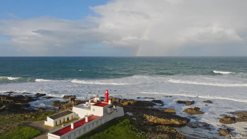 A scenic aerial view of a red and white lighthouse on a rocky coast as waves crash and a beautiful rainbow appears over the ocean