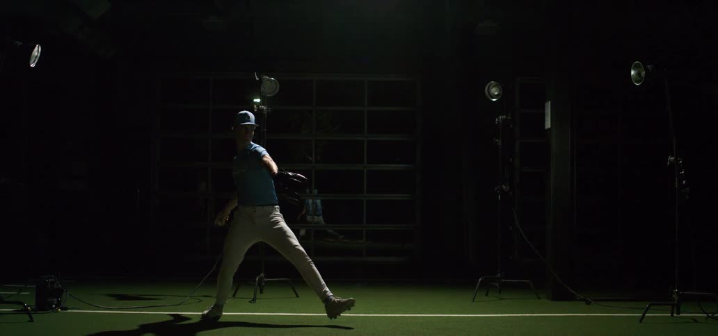 A baseball pitcher winds up and throws a pitch in dramatic slow motion as high-speed camera lights flash in a dark indoor training facility