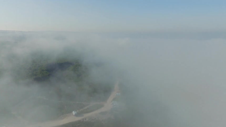 Ethereal aerial view pulling back through thick morning fog to reveal a wind farm and green landscape with the ocean on the horizon