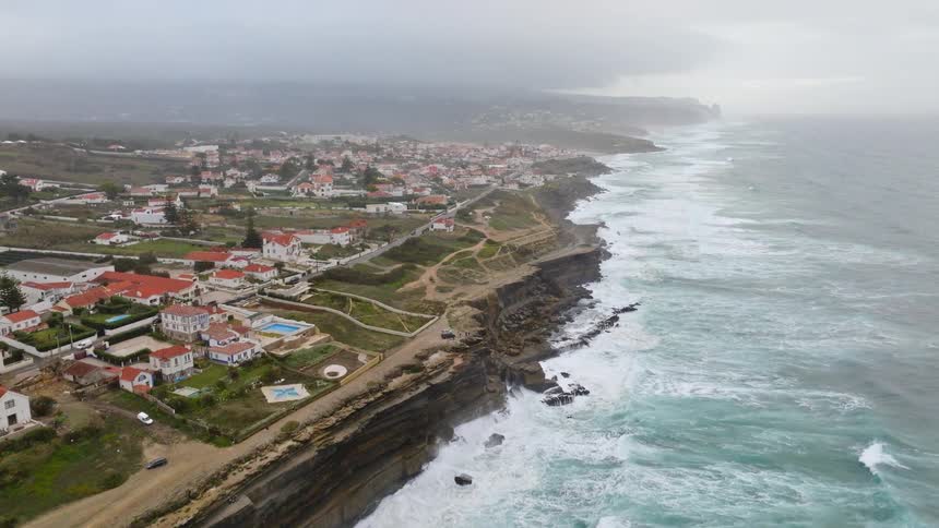 Aerial view of a coastal town on a cliffside with rough ocean waves crashing against the rocks on a stormy day