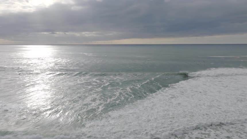 Slow-motion drone footage follows a surfer riding a perfect wave toward a rocky coastline under a dramatic, cloudy sky