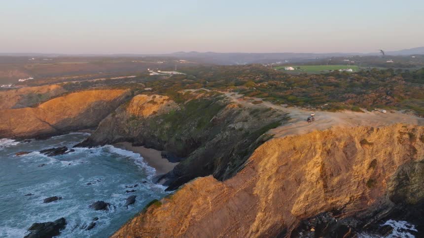 An aerial shot pulls back to reveal cars parked on a rugged, golden-lit cliff overlooking a beautiful coastline at sunset