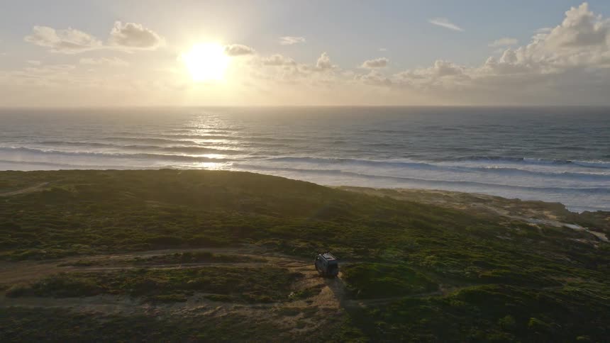 Aerial view of a camper van parked on a scenic, green coastline, overlooking the ocean at sunset