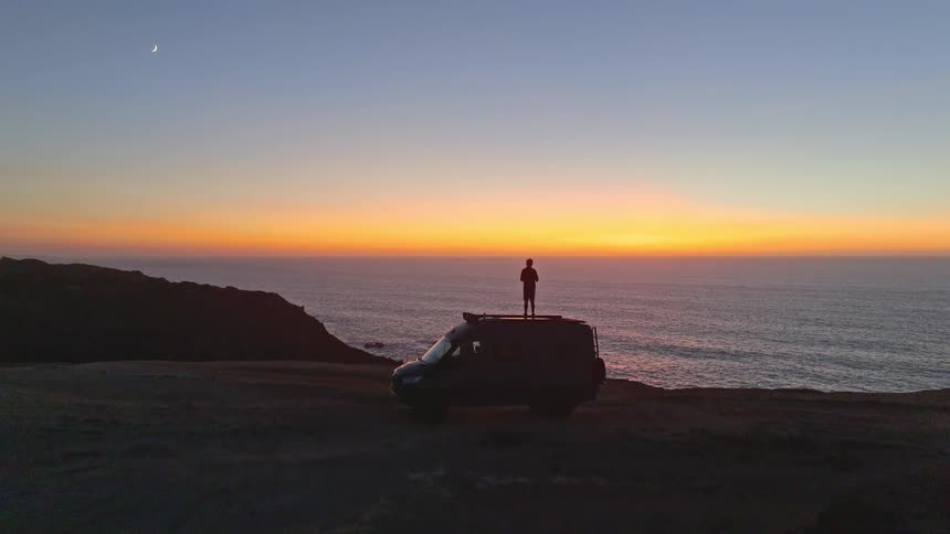 An aerial shot pushes in on a person silhouetted on their camper van, watching the vibrant sunset over the ocean from a cliffside with a crescent moon in the sky