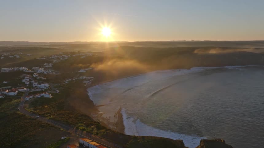 A stunning aerial view of a coastal town and beach as the sun rises, casting a golden glow over the misty ocean and rolling waves