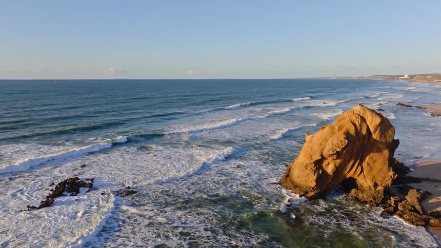 Aerial footage reveals a beautiful coastline with a large rock formation and waves crashing onto the sandy beach next to a coastal town during sunset
