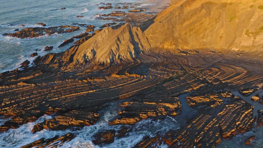 Stunning aerial shot pulls back from a detailed view of folded rock strata to reveal the magnificent and rugged coastline at golden hour