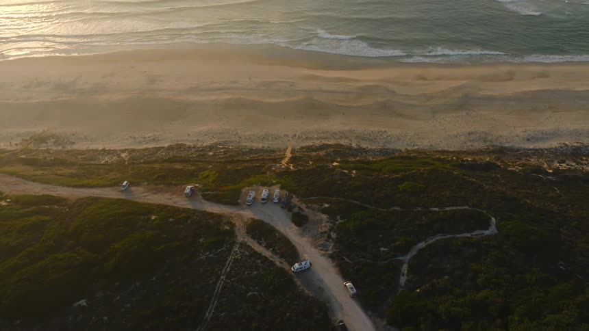 An aerial top-down view reveals camper vans parked on a dirt road by a sandy beach, tilting up to show the vast ocean at golden hour