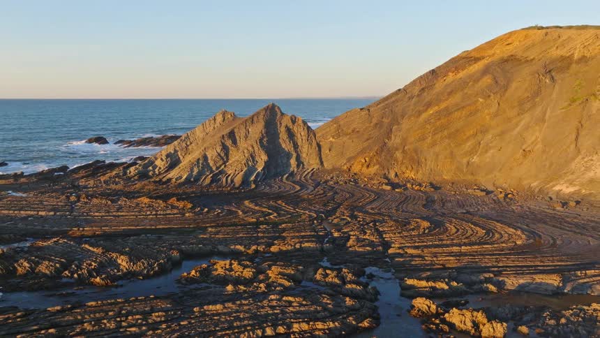 Aerial shot pulls back to reveal a stunning coastline with unique, layered rock formations glowing in the golden light of sunset