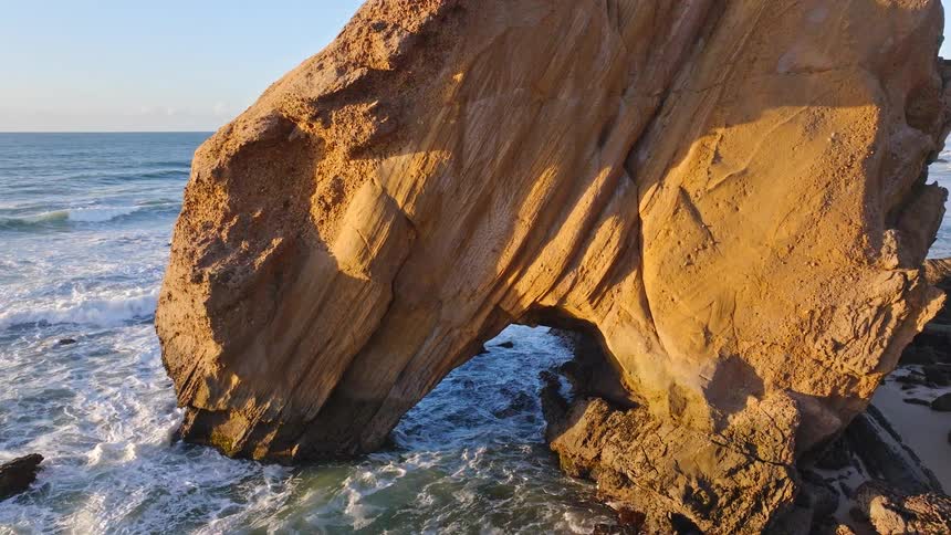 Golden hour light illuminates a massive rock arch on the coastline as ocean waves gently crash and flow through its opening