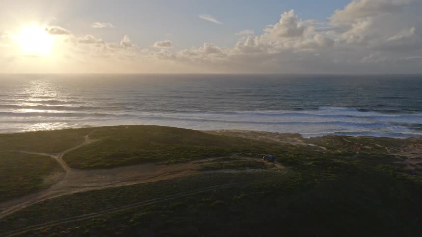 Panoramic aerial view of a car parked on a scenic coastal bluff overlooking the ocean at sunset