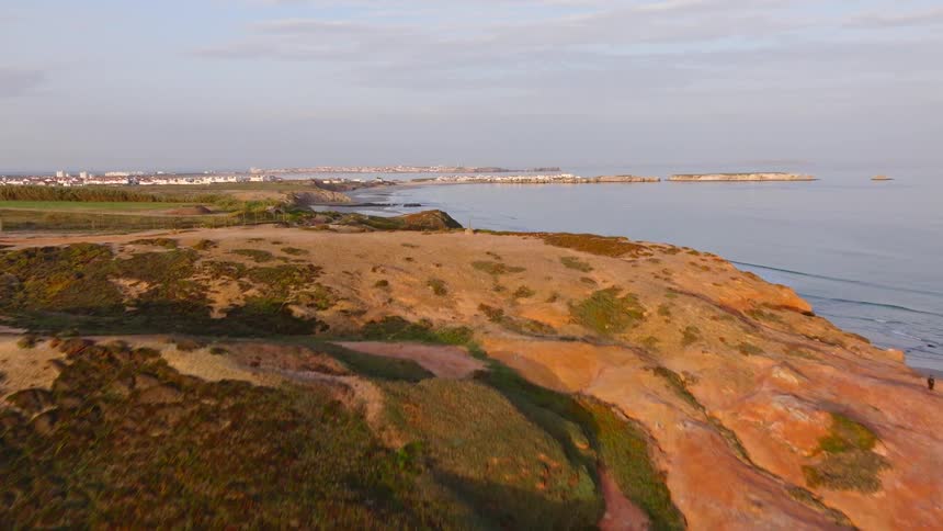 An aerial flyover of stunning red and green coastal cliffs reveals a vast sandy beach and the ocean at dawn