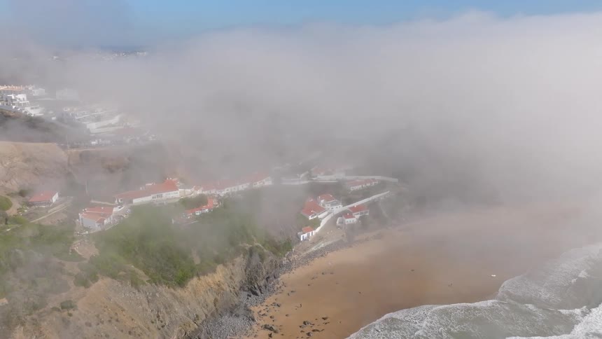 Aerial shot of a thick layer of fog rolling in from the ocean and covering a coastal village built on the cliffs