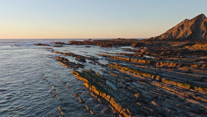 A slow aerial flight reveals a stunning rocky coastline with unique layered formations illuminated by the golden light of sunset