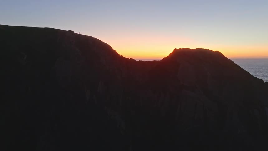 An aerial drone rises above a dark, silhouetted mountain cliff to reveal a beautiful, colorful ocean sunset on the horizon