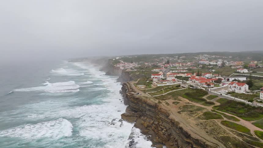 An epic aerial view ascends over a dramatic, rocky coastline revealing a picturesque seaside village on a foggy day
