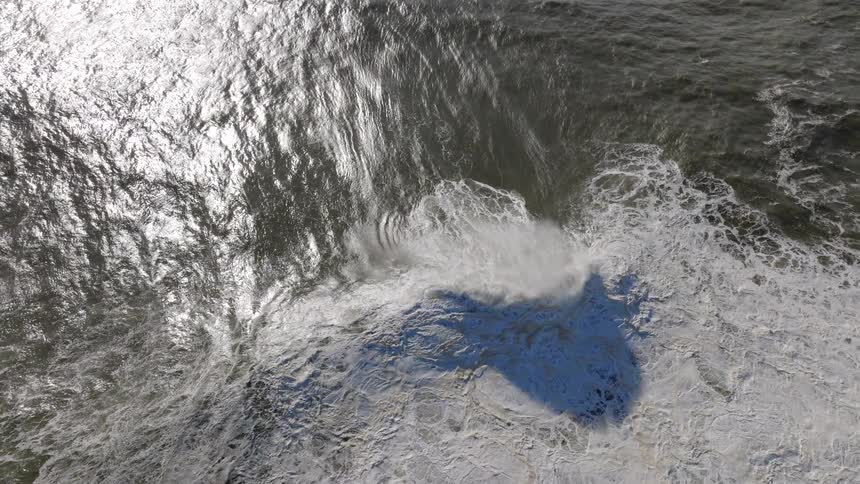 A dramatic slow-motion aerial view captures a powerful ocean wave as it curls and crashes into a sea of white foam