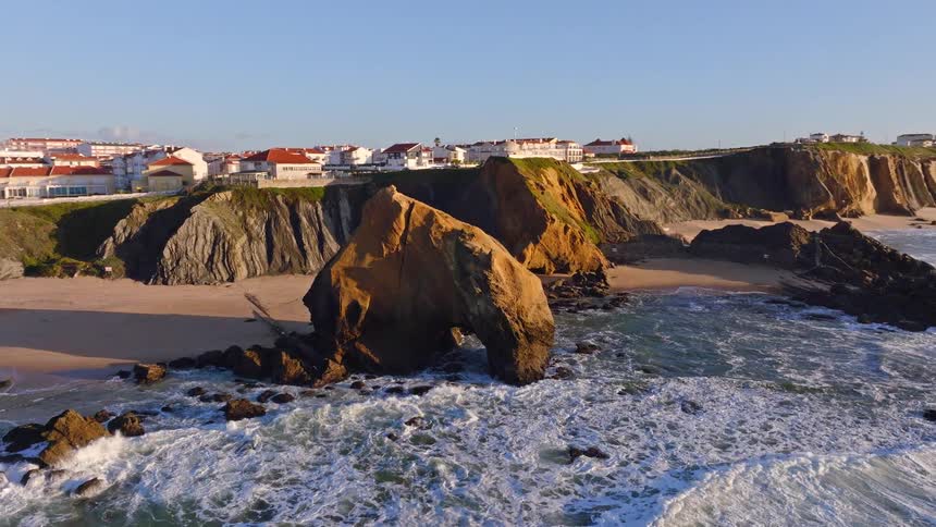 An aerial shot reveals the stunning Penedo do Guincho rock arch on a sandy beach in Santa Cruz, Portugal, as golden hour sunlight hits the cliffs