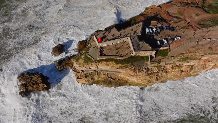 A stunning top-down aerial view captures the iconic Nazaré lighthouse and fort in Portugal, with tourists gathered to witness the immense power of crashing ocean waves