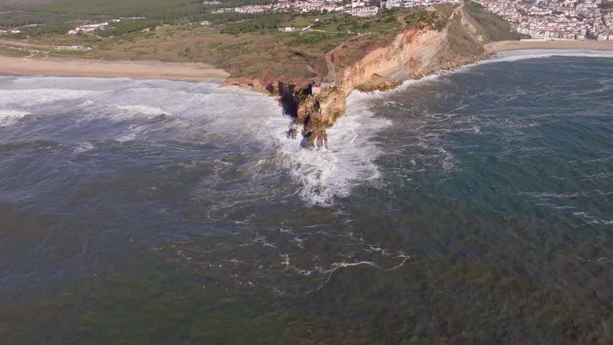An aerial pullback reveals the stunning and rugged coastline of Nazaré, Portugal, as powerful ocean waves crash against the cliffs