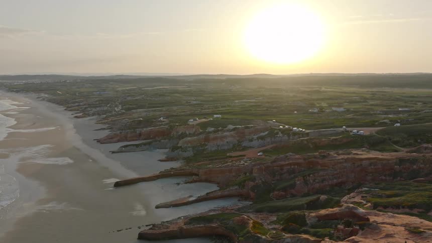 A serene aerial perspective reveals a vast coastline featuring reddish-brown cliffs, a sandy beach, and a golden sunset