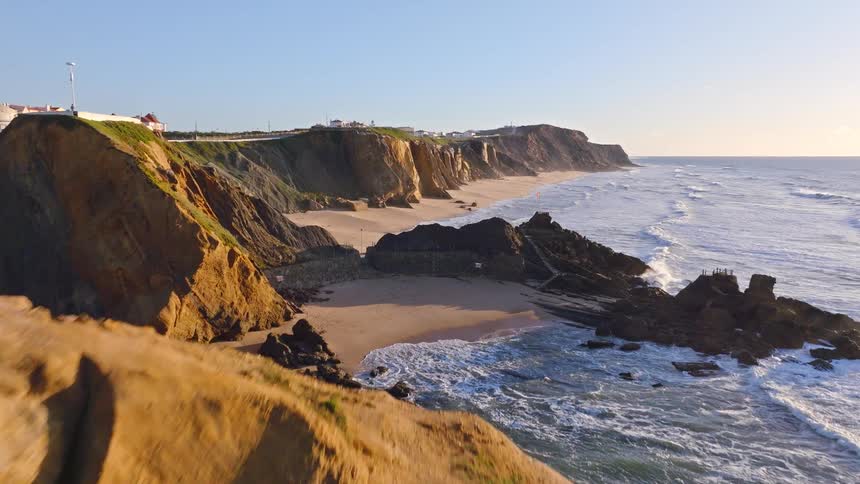 An aerial view glides over a magnificent rock arch and along a beautiful sandy coastline with dramatic cliffs during a golden sunset