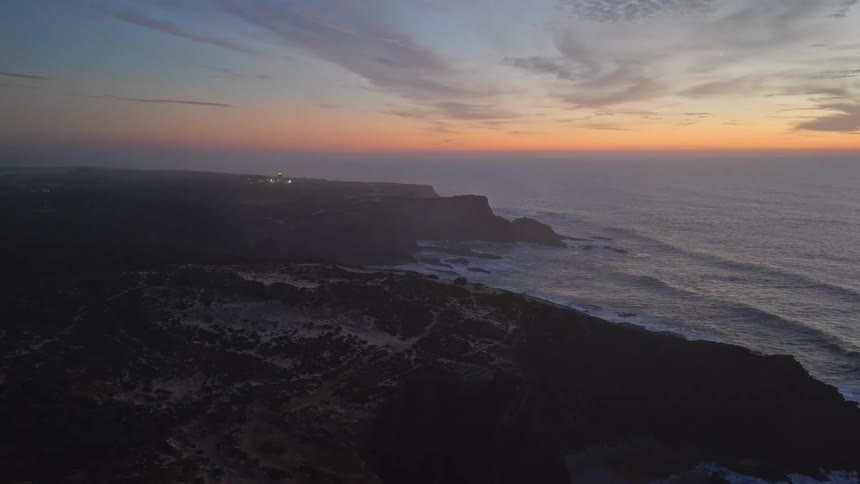 An aerial view shows a beautiful coastline with a lighthouse in the background at sunset, as the camera slowly pulls back