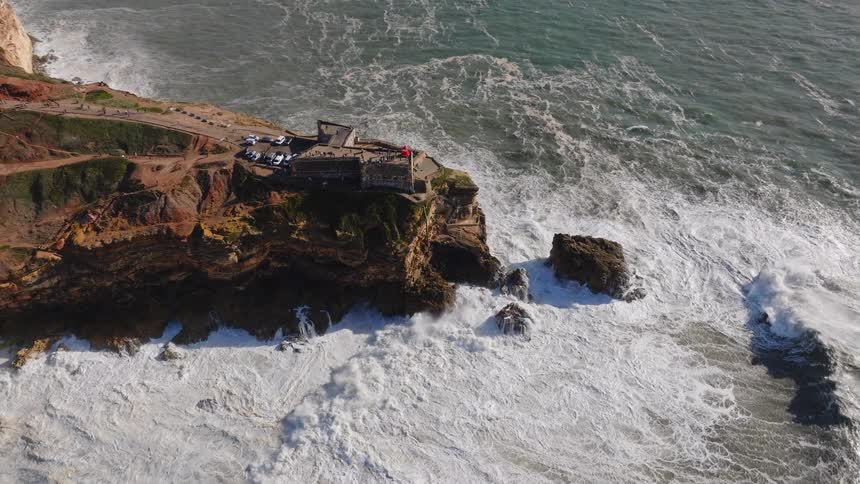 A dramatic aerial view pans out from the iconic Nazaré lighthouse as massive, foamy ocean waves pound against the rocky cliffs of Portugal's coastline