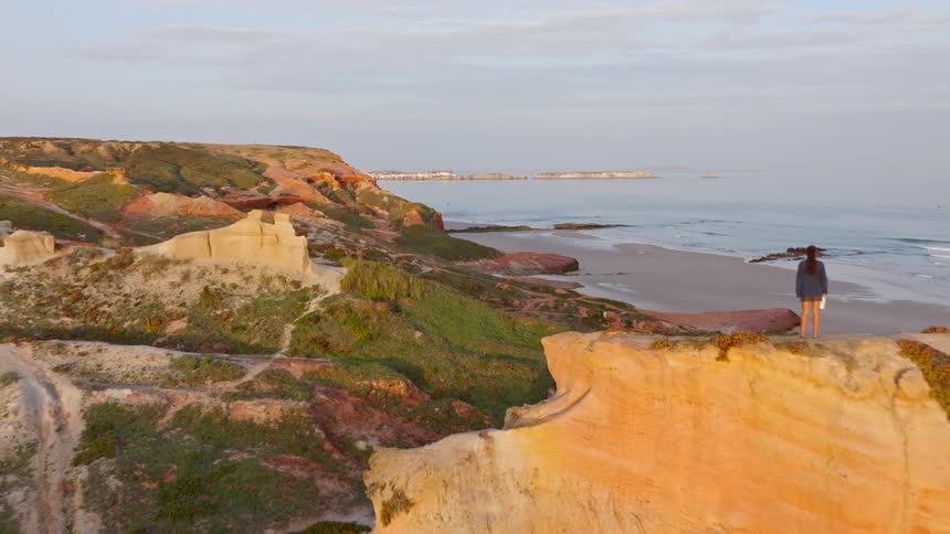 A stunning aerial view pulls back to reveal a woman standing atop colorful cliffs overlooking the ocean at sunrise, with a camper van parked nearby