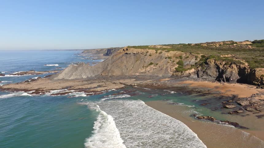 A stunning aerial view reveals a rugged and scenic coastline with dramatic cliffs, a sandy beach, and beautiful turquoise ocean waves
