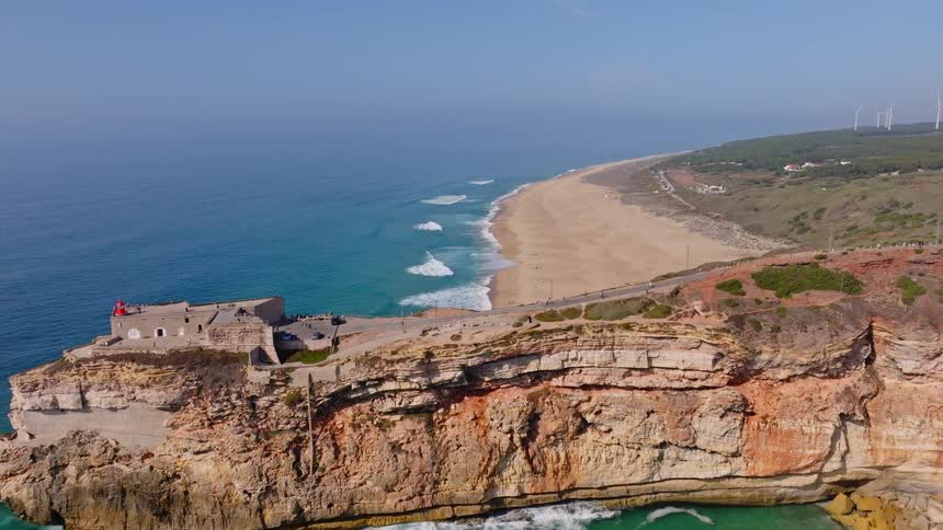 A stunning aerial pullback reveals the historic Fort of So Miguel Arcanjo on a dramatic cliff overlooking the vast Praia do Norte beach in Nazaré, Portugal