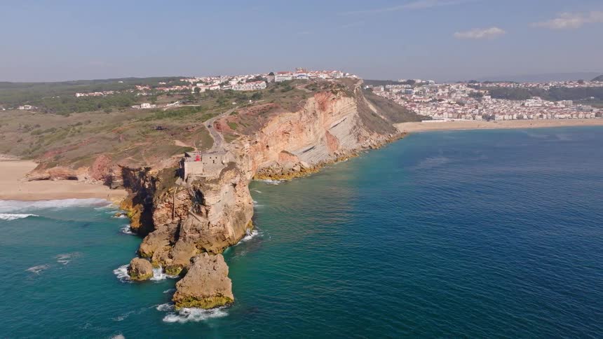 Aerial view of the stunning coastline of Nazaré, Portugal, with the São Miguel Arcanjo Fort perched on a cliff overlooking the turquoise ocean and sandy beaches