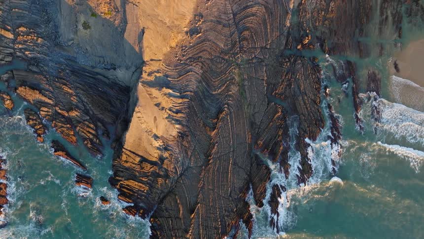 A slow ascending top-down aerial shot reveals the incredible patterns of a folded rock formation on the coast as ocean waves crash against the shore during a golden sunset