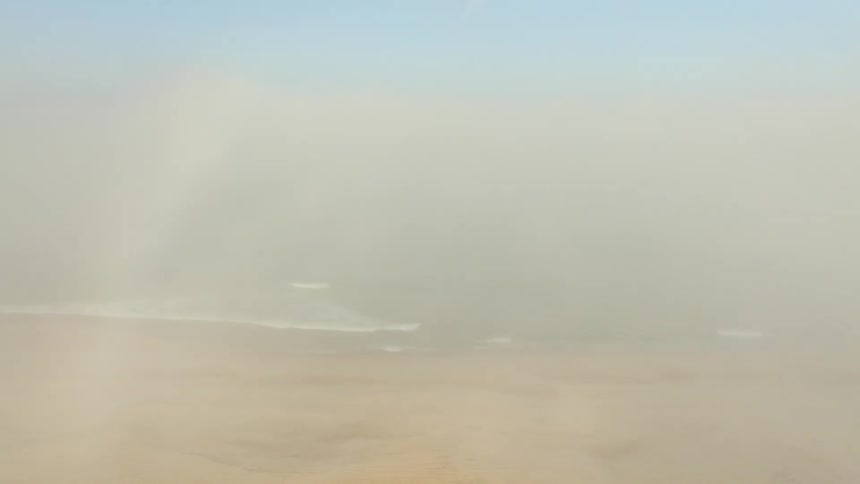 An ethereal aerial shot flying through a thick layer of fog, gradually revealing a vast sandy beach and the gentle waves of the ocean below