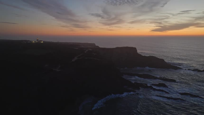 A serene aerial shot captures a lighthouse on a rugged coastline as its light beam sweeps across the landscape and sea after sunset