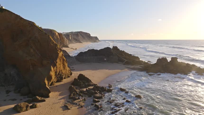 An expansive aerial pullback reveals the breathtaking beauty of the Portuguese coastline at Praia de Santa Cruz during a golden sunset, featuring cliffs, waves, and a national flag