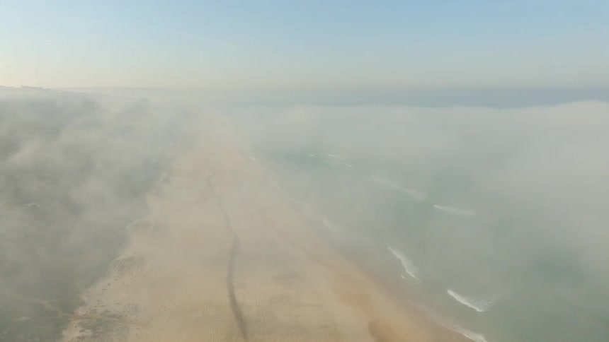 An ethereal aerial shot ascends through a thick blanket of fog to reveal a stunning sandy beach and the vast ocean coastline