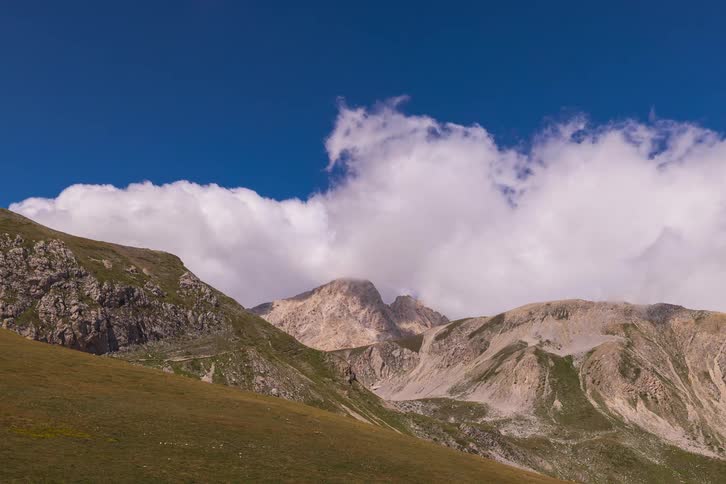 A scenic timelapse of fluffy white clouds moving over a majestic, rocky mountain range with green hills in the foreground
