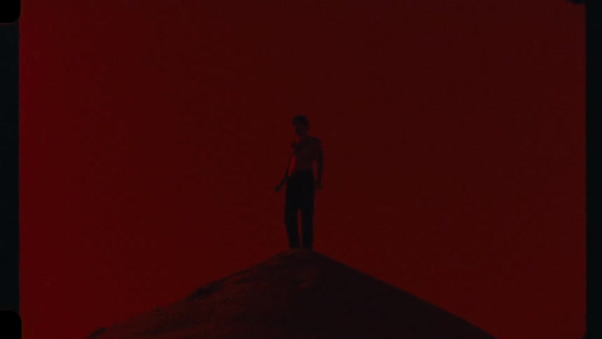 A mysterious man is silhouetted on a mountaintop in a desolate, otherworldly landscape with a deep red sky