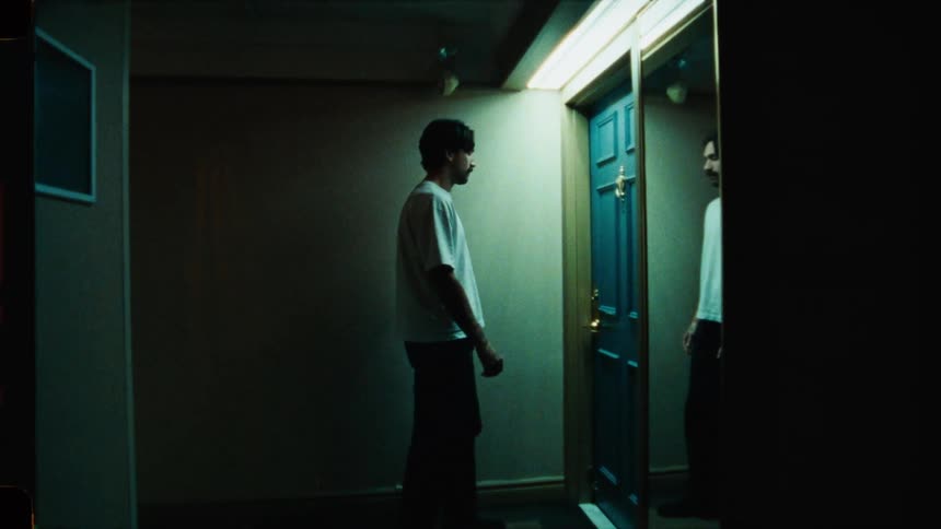 A man slowly walks down a dark, cinematic hallway, stopping to see his reflection in a mirror before reaching for the doorknob of a blue door