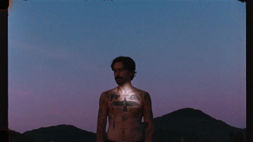 An artistic slow zoom on a pensive, shirtless man with tattoos standing against a mountain range at dusk